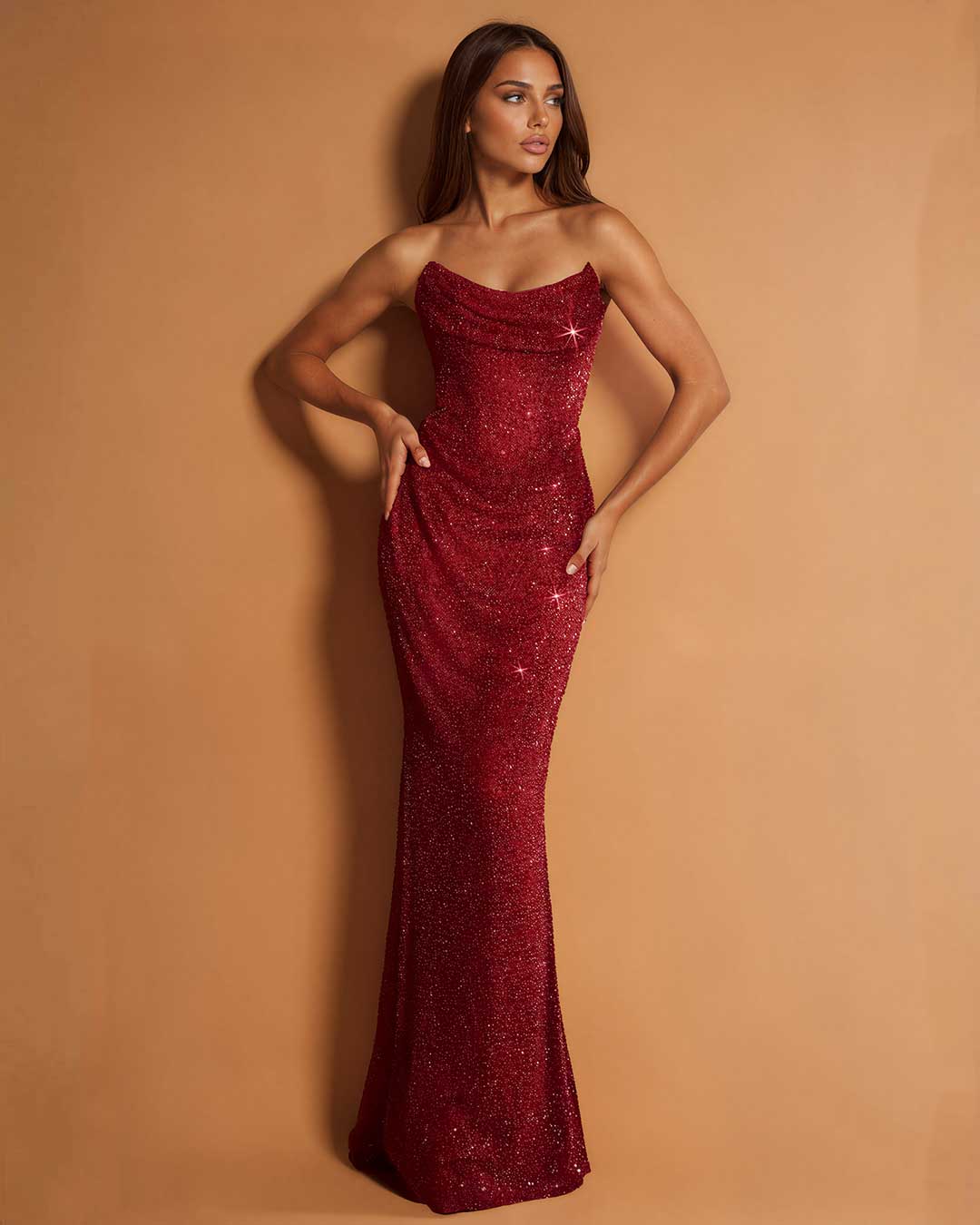 A woman modeling a strapless, floor-length mermaid gown covered in shimmering red sequins, posed against a solid warm tan studio backdrop.