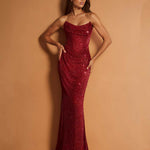 A woman modeling a strapless, floor-length mermaid gown covered in shimmering red sequins, posed against a solid warm tan studio backdrop.