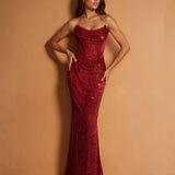 A woman modeling a strapless, floor-length mermaid gown covered in shimmering red sequins, posed against a solid warm tan studio backdrop.
