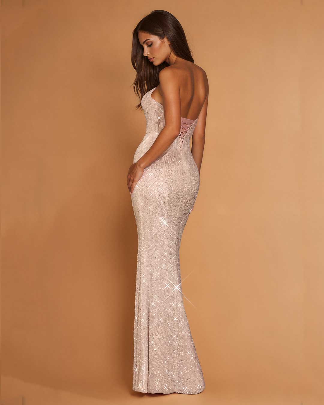 A woman modeling a sparkling champagne sequined evening gown with a backless open back and lace-up detail, posed against a solid warm tan studio backdrop.