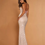 A woman modeling a sparkling champagne sequined evening gown with a backless open back and lace-up detail, posed against a solid warm tan studio backdrop.