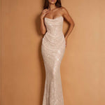 A woman modeling a strapless, floor-length mermaid gown covered in shimmering sequins, posed against a warm tan studio backdrop.