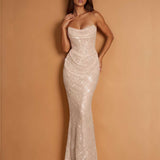 A woman modeling a strapless, floor-length mermaid gown covered in shimmering sequins, posed against a warm tan studio backdrop.