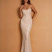 A woman modeling a strapless, floor-length mermaid gown covered in shimmering sequins, posed against a warm tan studio backdrop.