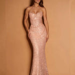 A woman modeling a strapless, sweetheart-neckline mermaid gown covered in shimmering rose-gold sequins, set against a solid warm tan studio backdrop.
