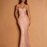 A woman modeling a strapless, sweetheart-neckline mermaid gown covered in shimmering rose-gold sequins, set against a solid warm tan studio backdrop.