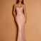 A woman modeling a strapless, sweetheart-neckline mermaid gown covered in shimmering rose-gold sequins, set against a solid warm tan studio backdrop.