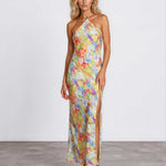 A woman modeling a halter-neck, floor-length maxi dress with a vibrant multicolor floral print on a light yellow base, standing against a plain white studio wall.