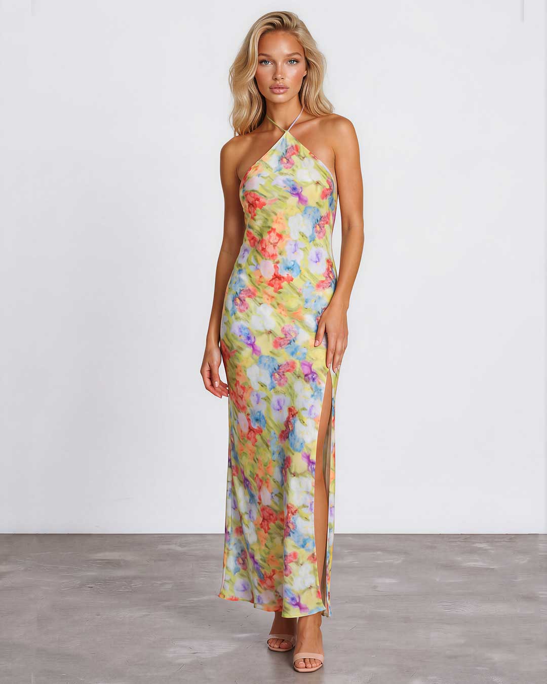 A woman modeling a halter-neck, floor-length maxi dress with a vibrant multicolor floral print on a light yellow base, standing against a plain white studio wall.