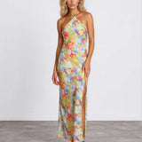 A woman modeling a halter-neck, floor-length maxi dress with a vibrant multicolor floral print on a light yellow base, standing against a plain white studio wall.