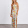 A woman modeling a halter-neck, floor-length maxi dress with a vibrant multicolor floral print on a light yellow base, standing against a plain white studio wall.