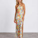 A woman modeling a halter-neck, floor-length maxi dress with a vibrant multicolor floral print on a light yellow base, standing against a plain white studio wall.
