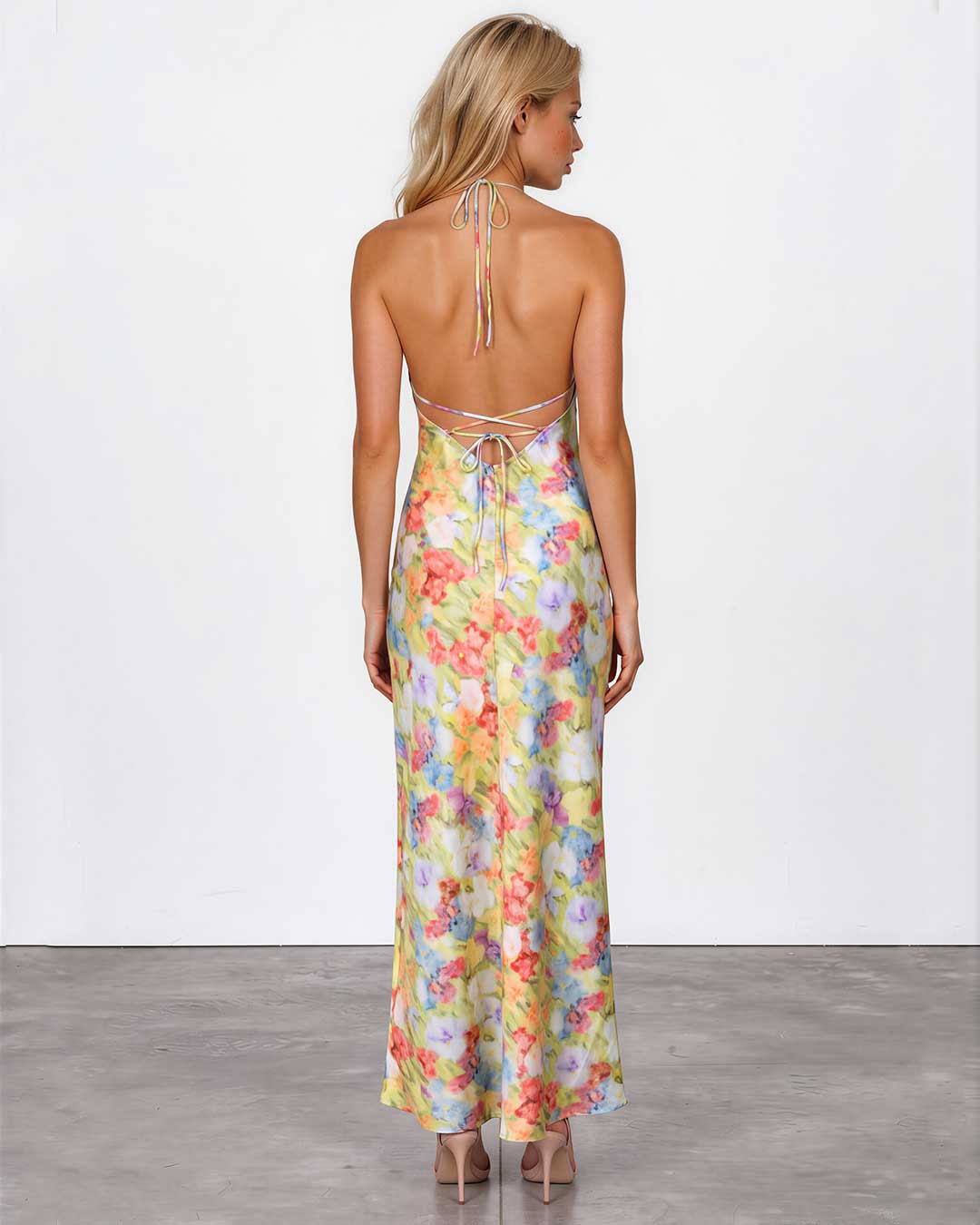 A woman stands with her back to the camera wearing a sleeveless, open-back maxi dress with a pastel multicolor floral print and tie-shoulder straps, paired with nude heels against a white studio wall and gray floor.
