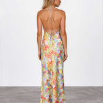 A woman stands with her back to the camera wearing a sleeveless, open-back maxi dress with a pastel multicolor floral print and tie-shoulder straps, paired with nude heels against a white studio wall and gray floor.