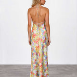 A woman stands with her back to the camera wearing a sleeveless, open-back maxi dress with a pastel multicolor floral print and tie-shoulder straps, paired with nude heels against a white studio wall and gray floor.