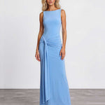 A woman modeling a sleeveless light blue maxi dress with a draped, gathered waist detail, standing against a plain white studio wall and gray floor.