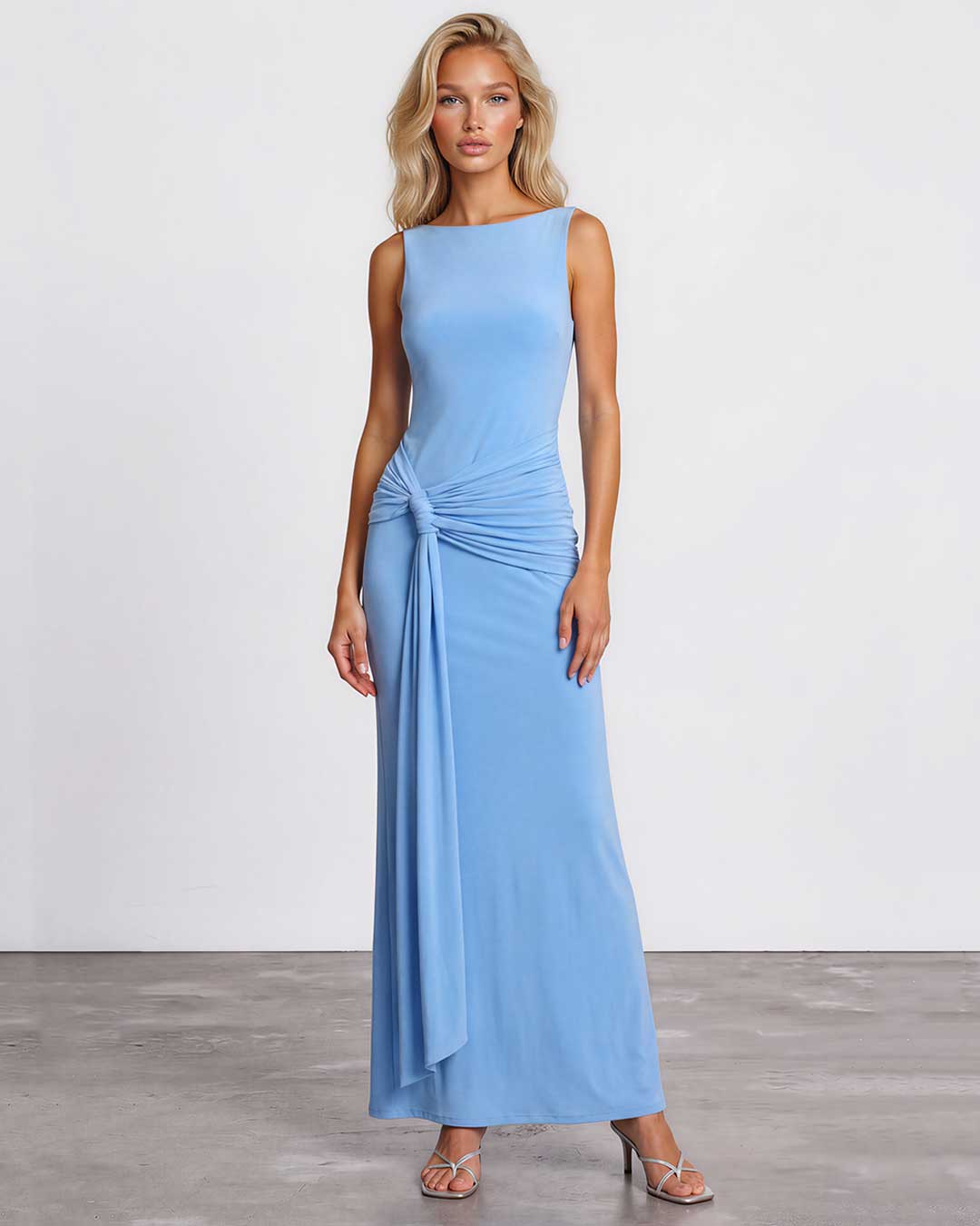A woman modeling a sleeveless light blue maxi dress with a draped, gathered waist detail, standing against a plain white studio wall and gray floor.