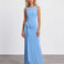 A woman modeling a sleeveless light blue maxi dress with a draped, gathered waist detail, standing against a plain white studio wall and gray floor.