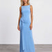 A woman modeling a sleeveless light blue maxi dress with a draped, gathered waist detail, standing against a plain white studio wall and gray floor.