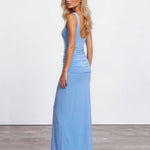 A woman modeling a light blue sleeveless maxi dress with a ruched bodice and backless open back, paired with clear high-heeled sandals, set against a plain white studio wall and gray floor.