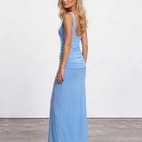A woman modeling a light blue sleeveless maxi dress with a ruched bodice and backless open back, paired with clear high-heeled sandals, set against a plain white studio wall and gray floor.