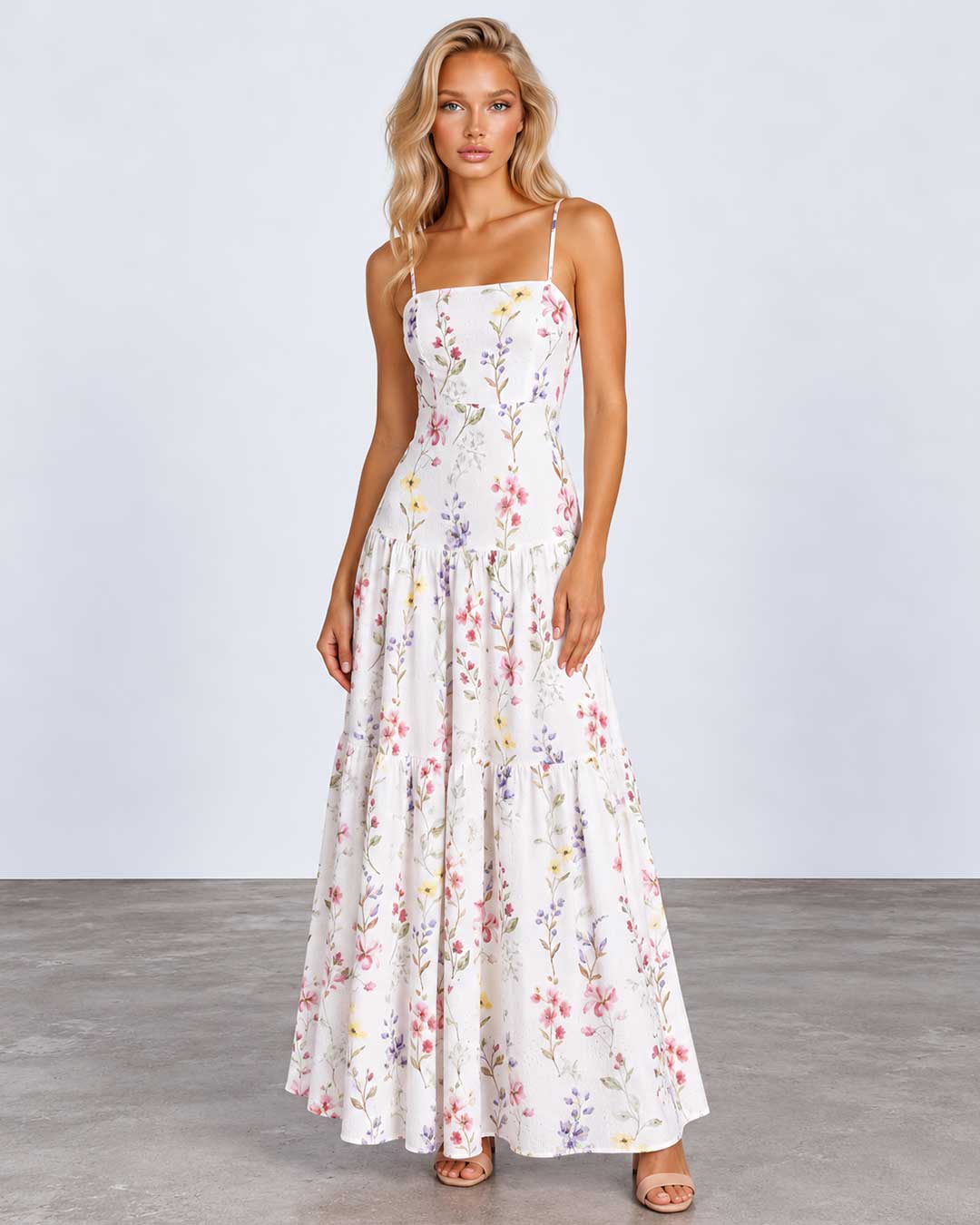 A woman modeling a sleeveless, tiered maxi dress with a delicate multicolor floral print on a white background, standing against a light gray studio backdrop.