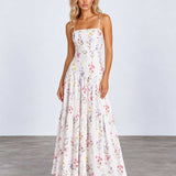 A woman modeling a sleeveless, tiered maxi dress with a delicate multicolor floral print on a white background, standing against a light gray studio backdrop.
