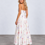 A woman modeling a white spaghetti-strap maxi dress with a colorful pastel floral print, shown from the back against a light gray studio background.