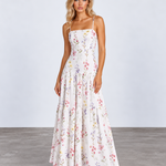 A woman modeling a white spaghetti-strap maxi dress with a colorful pastel floral print, standing against a light gray studio background.
