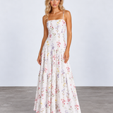 A woman modeling a white spaghetti-strap maxi dress with a colorful pastel floral print, standing against a light gray studio background.
