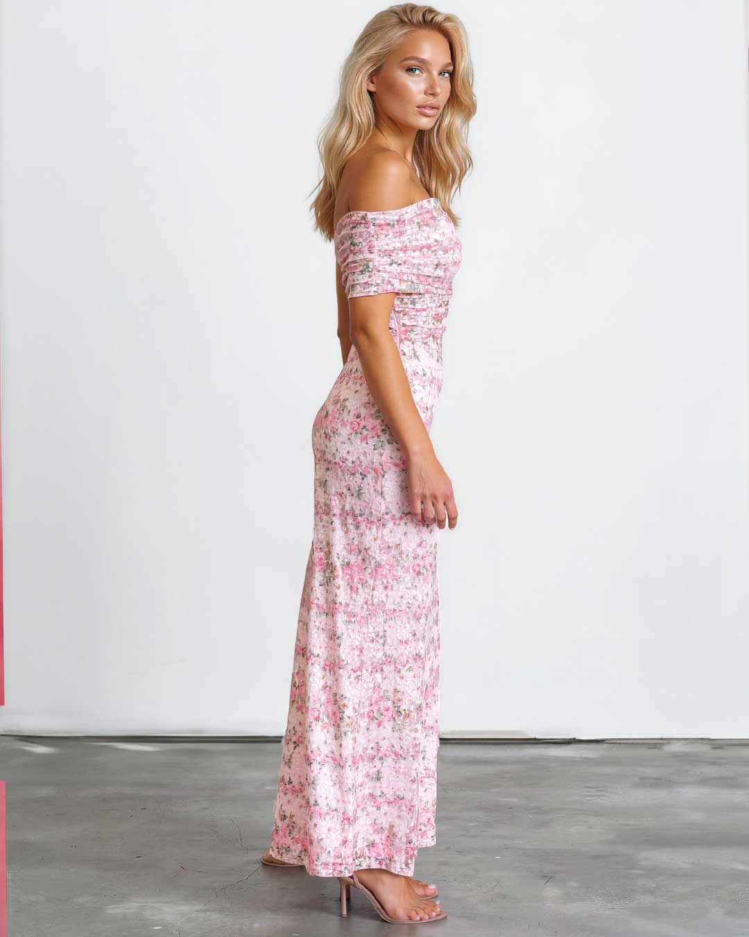 A woman modeling an off-the-shoulder maxi dress with a pink and white floral pattern, standing in a studio with a white wall and gray floor.