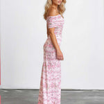 A woman modeling an off-the-shoulder maxi dress with a pink and white floral pattern, standing in a studio with a white wall and gray floor.