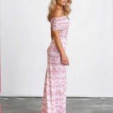 A woman modeling an off-the-shoulder maxi dress with a pink and white floral pattern, standing in a studio with a white wall and gray floor.