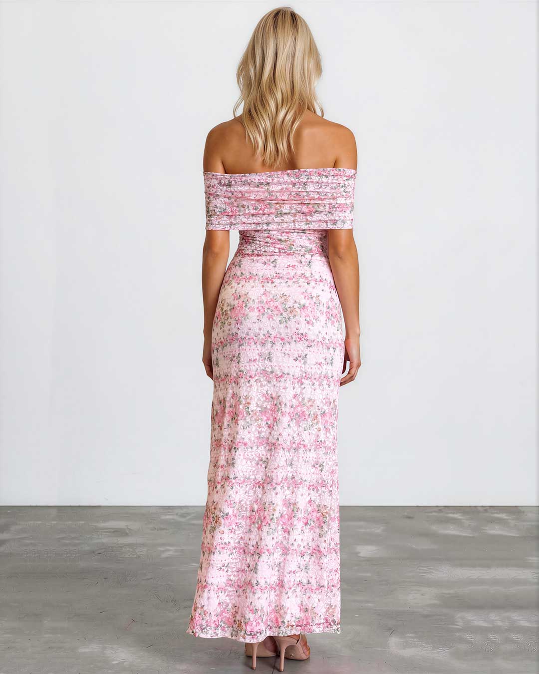 A woman viewed from behind wearing a long off-the-shoulder maxi dress with a pink and white floral pattern, standing against a plain white wall on a gray concrete floor.