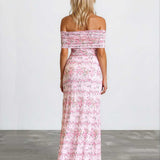 A woman viewed from behind wearing a long off-the-shoulder maxi dress with a pink and white floral pattern, standing against a plain white wall on a gray concrete floor.