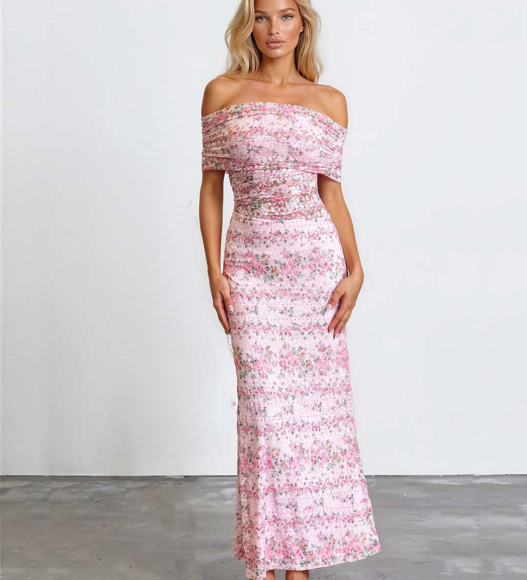 A woman modeling an off-the-shoulder, floor-length pink floral dress against a plain white studio wall and gray floor.