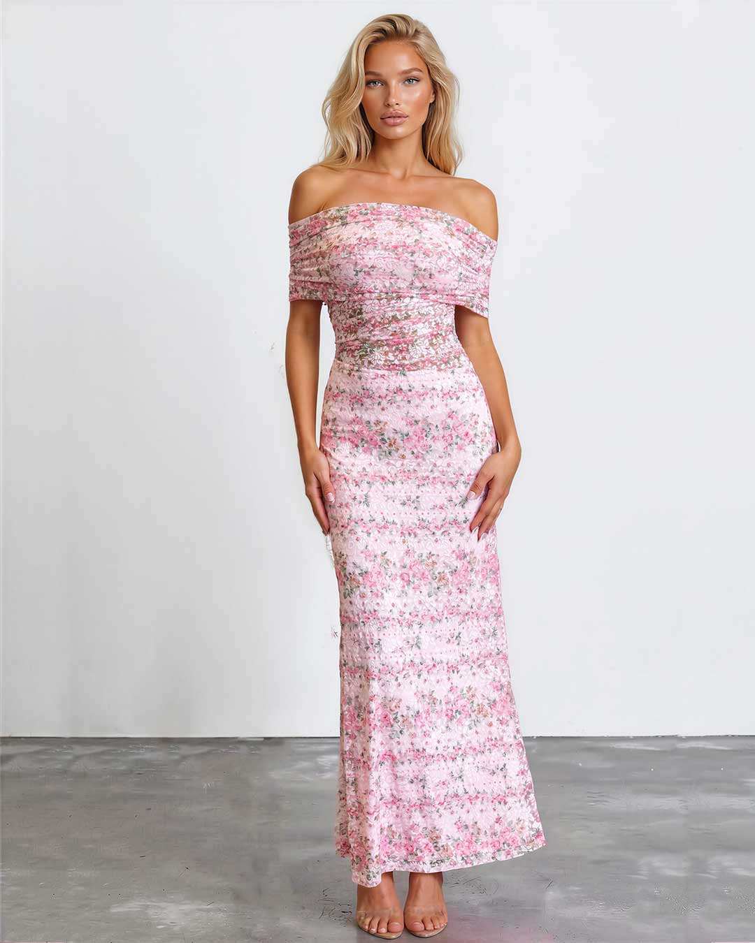 A woman modeling an off-the-shoulder, floor-length pink floral dress against a plain white studio wall and gray floor.