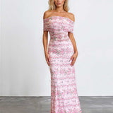 A woman modeling an off-the-shoulder, floor-length pink floral dress against a plain white studio wall and gray floor.
