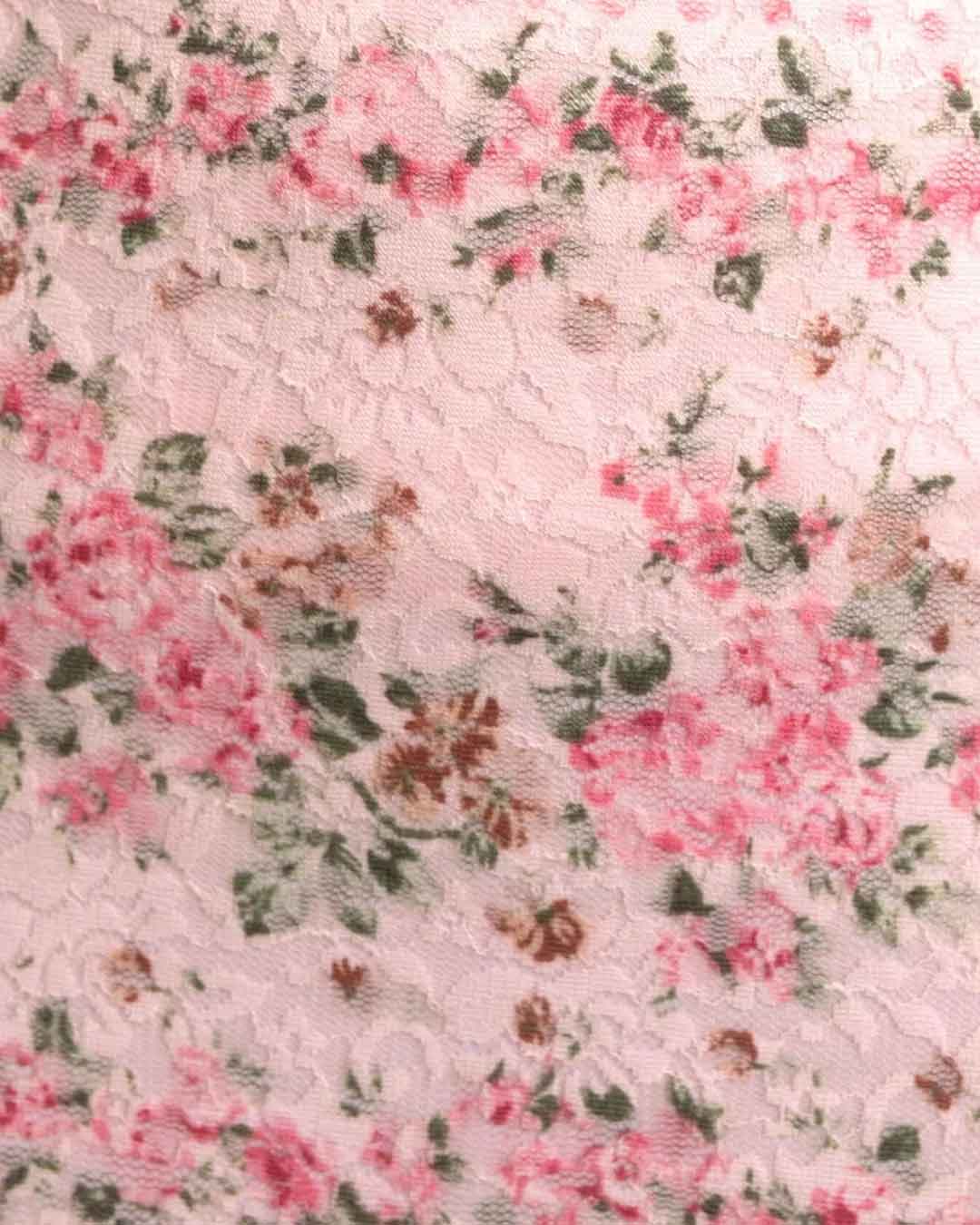 A close-up of a light pink floral lace fabric with pink roses, green leaves, and brown accents.