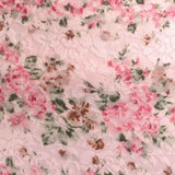 A close-up of a light pink floral lace fabric with pink roses, green leaves, and brown accents.