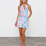 A woman modeling a light blue halter mini dress with a pink and green floral print, standing against a white wall and gray concrete floor.
