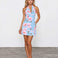 A woman modeling a light blue halter mini dress with a pink and green floral print, standing against a white wall and gray concrete floor.