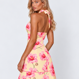 A woman modeling a pastel yellow halter mini dress with a pink floral print, shown from the back against a light gray studio background.