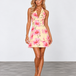 A blonde model wearing a sleeveless halter mini dress with a pink floral print on a pale yellow background, standing against a light gray studio wall.