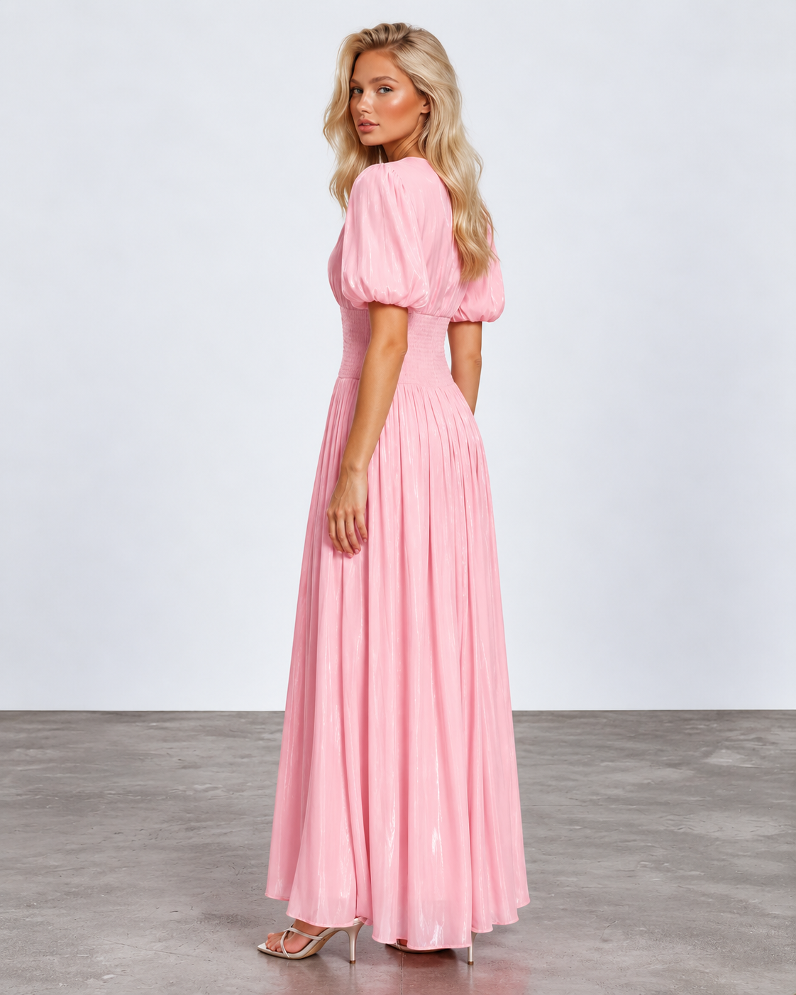 A woman modeling a long pink maxi dress with puff sleeves and a pleated skirt, standing in a studio with a light gray wall and concrete floor.