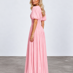 A woman modeling a long pink maxi dress with puff sleeves and a pleated skirt, standing in a studio with a light gray wall and concrete floor.