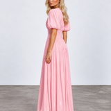 A woman modeling a long pink maxi dress with puff sleeves and a pleated skirt, standing in a studio with a light gray wall and concrete floor.