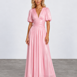 A woman modeling a floor-length metallic pink gown with puff sleeves, a deep V-neckline, and a gathered waist, photographed against a light gray studio backdrop.