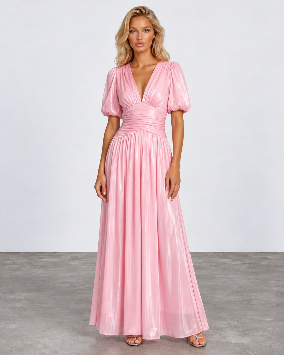 A woman modeling a floor-length metallic pink gown with puff sleeves, a deep V-neckline, and a gathered waist, photographed against a light gray studio backdrop.