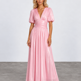 A woman modeling a floor-length metallic pink gown with puff sleeves, a deep V-neckline, and a gathered waist, photographed against a light gray studio backdrop.